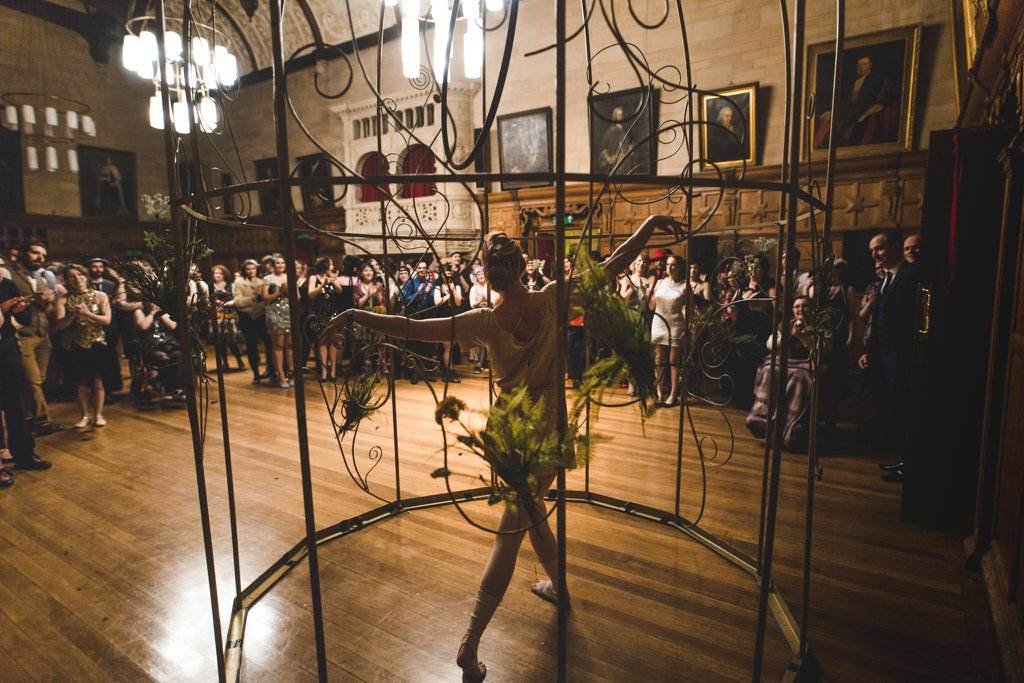 An aerial performer stands in a giant gold birdcage in front of a crowd