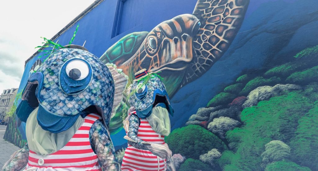 two performers dressed as reverse mermaids infront of a painting of some fish