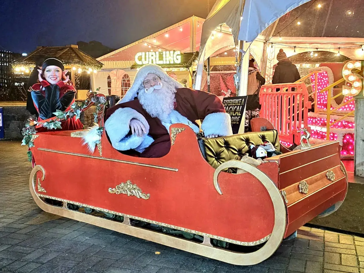 A santa act featuring santa sitting in a mobile sleigh next to his elf helper who is leaning on the back and smiling.