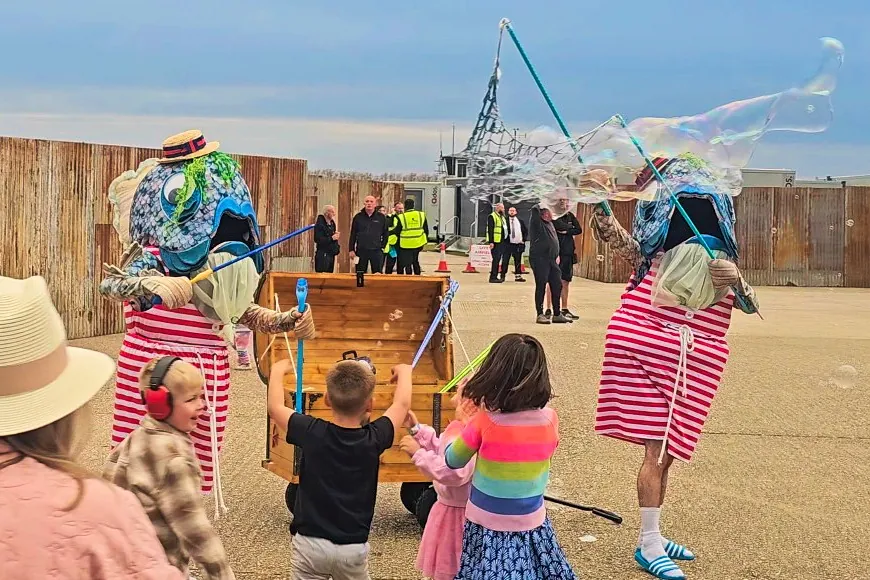 Children enjoying seaside entertainment featuring bubbles created by two performers in comedy fish outfits.