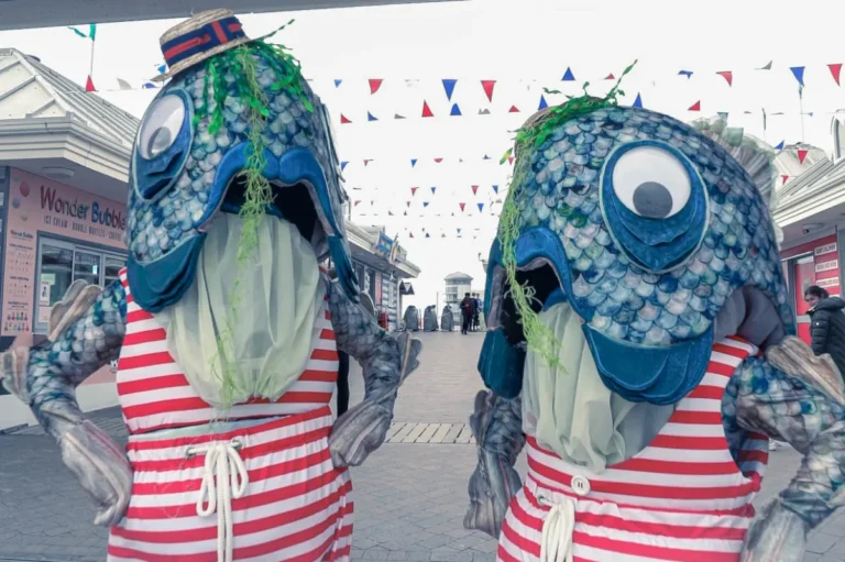 Two comedy performers in fish outfits standing with hands on hips in a seaside environment.