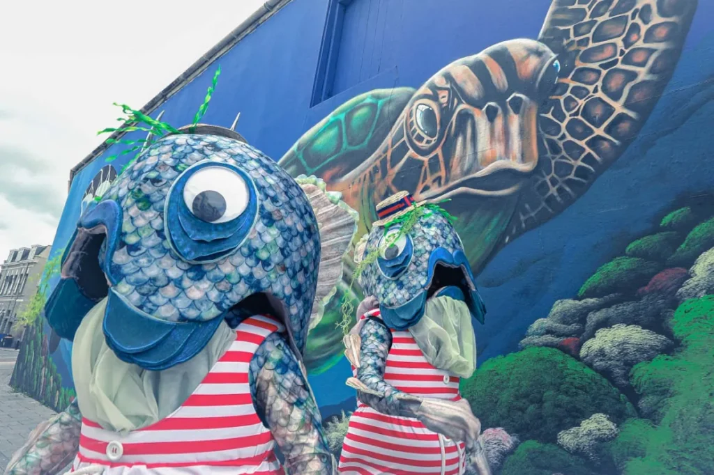 Two performers in comedy fish outfits strutting past a large sea themed mural.