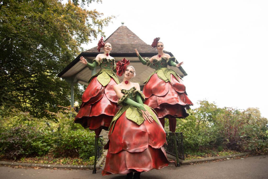 Three female performers in rose themed costumes, two of them are stood on stilts.